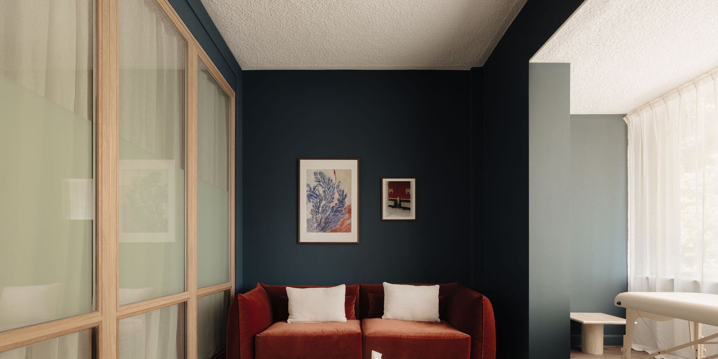 A quiet consulting room with a deep blue wall, a rust-red sofa with cream cushions, a framed picture and a wood-framed glass partition