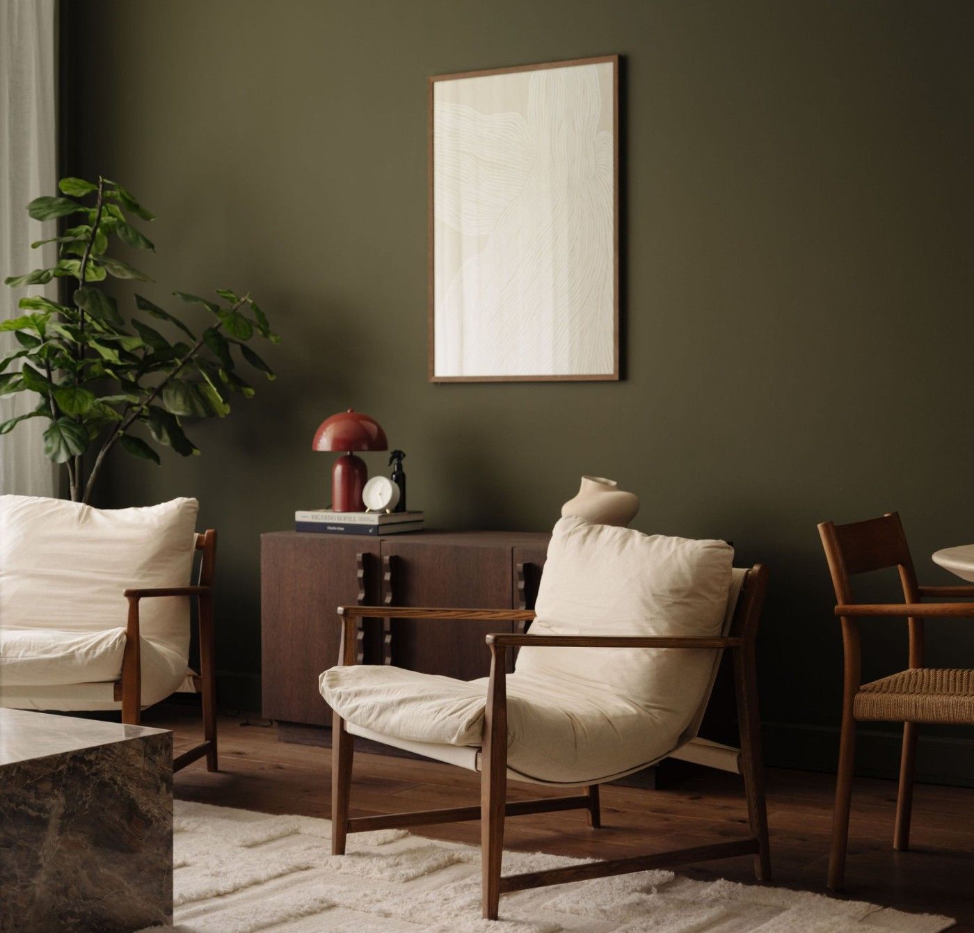 A quiet room with an olive green wall, a framed print, a cream armchair with a wooden frame, a dark wood sideboard with a red lamp, and a fiddle leaf plant