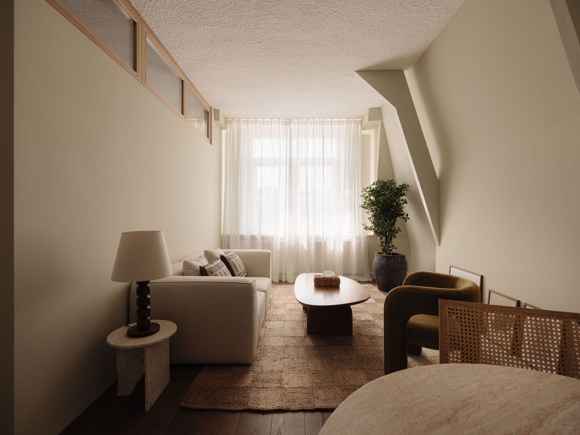 A calm room with a sofa, a low wooden table, a plant and sheer curtains at the window