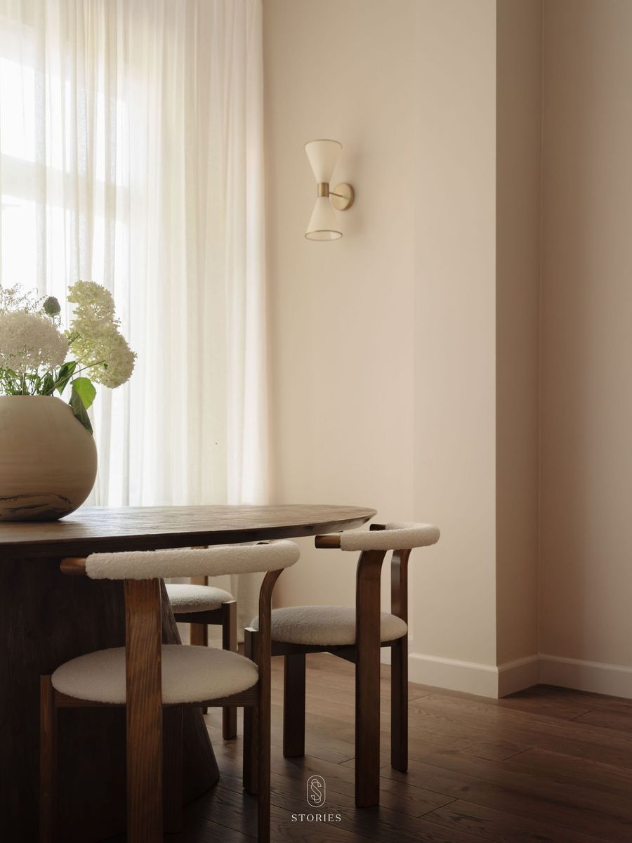 A round wooden table with curved chairs next to a window with sheer curtains, and a wall light
