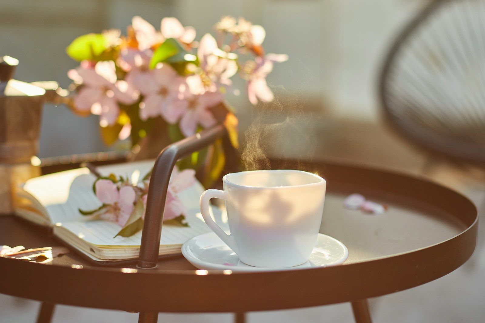 A cup of coffee on a tray next to an open book and pink spring blossom, in soft sunlight